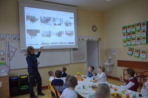 Policjantka prowadzi zajęcia, stoi przy tablicy, w tle wyświetlana jest prezentacja multimedialna.