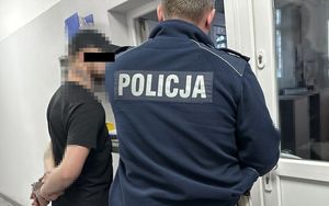 Policjanci stoją przy oknie dyżurnego, gdzie prowadzone jest badanie trzeźwości, mężczyzna ma kajdanki założone na ręce z tyłu.