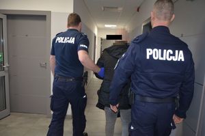 Policjanci prowadzą zatrzymanego mężczyznę korytarzem komendy, w kierunku wyjścia.