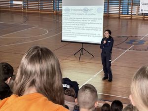 Policjantka prowadzi spotkanie z młodzieżą, dzieci siedzą na trybunach na hali gimnastycznej.