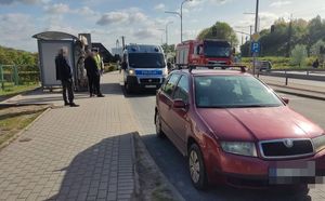 Na pierwszym planie widoczna jest czerwona skoda a za nią stoi radiowóz, obok niego po prawej stronie zdjęcia stoi wóz straży pożarnej. Na przystanku obok radiowozu stoi policjant, który rozmawia z jakimś mężczyzną.