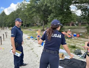 W tle zdjęcia widoczne jest jezioro oraz plaża, a na niej osoby, które wypoczywają. Na pierwszym planie jest policjantka, która wręcza dziecku jakiś drobny upominek, a przy niej stoi dzielnicowy.