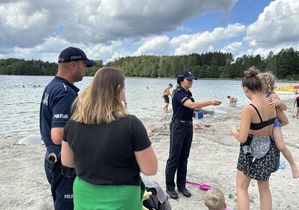 W tle zdjęcia widoczne jest jezioro oraz plaża, a na niej osoby, które wypoczywają. Na pierwszym planie jest policjantka, która wręcza dziecku trzymanemu na rękach przez kobietę, jakiś drobny upominek, a przy niej stoi dzielnicowy , a przed nim stoi także inna kobieta.