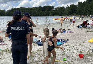 W tle zdjęcia widoczne jest jezioro oraz plaża, a na niej osoby, które wypoczywają. Na pierwszym planie jest policjantka, która stoi obok dzielnicowego wręczającemu dziecku jakiś drobny upominek.