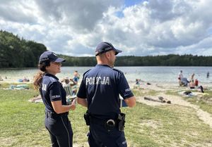 Policjanci wchodzą na plażę nad jeziorem, rozglądają się, w tle widoczni turyści oraz jezioro.