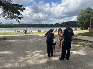 Policjanci przed wejściem na plażę rozmawiają z kobietą i małym dzieckiem, policjantka pochyla się do dziecka i wręcza mu upominek. Obok policjantki stoi dzielnicowy.