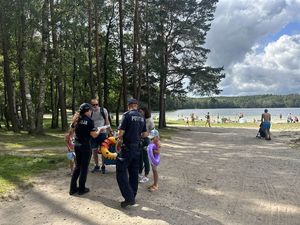 Policjanci rozmawiają z turystami opuszczającymi plażę. Turystami są dwie osoby dorosłe (kobieta i mężczyzna), niosący sprzęty pływackie (dmuchane koła i inne zabawki) oraz dwoje dzieci.