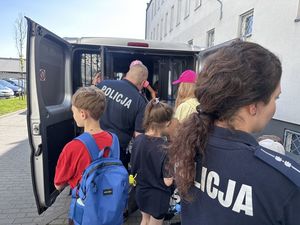 Dzieci z policjantką i innym policjantem stoją przed radiowozem typu bus w kolorze szarym. Tylne drzwi pojazdu są otwarte a dzieci wchodzą do środka.