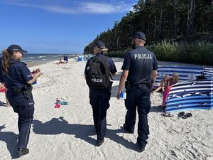 Troje policjantów idzie plażą, w tle widoczni turyści, którzy wypoczywają na kocach lub są za parawanami.