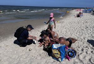 Policjantka przykucnęła przy rodzinie wypoczywającej z dziećmi na plaży. Rozmawia z turystami, dzieciom wręcza opaski niezgubki.