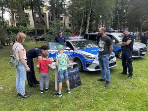 Dwójka dzieci podeszła do policjantki stojącej przy niewielkich rozmiarów stoliku. Wokół dzieci stoją dorośli opiekunowie po prawej mężczyzna, po lewej stronie kobieta. W tle stoją także dwaj policjanci. Policjantka przy stoliku pochyla się do dzieci, które do niej podeszły i coś im prezentuje.