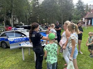 Policjantka rozmawia z grupą dzieci i ich opiekunami. W tle stoi policyjny radiowóz oraz policjant.