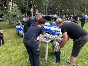 Policjantka z dzielnicowym wykonują małemu dziecku pamiątkową daktyloskopię. Obok dziecka stoi rodzic.