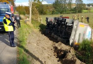 Policjant stoi na skraju jedni, coś pisze, przed nim na boku leży wywrócony pojazd ciężarowy.