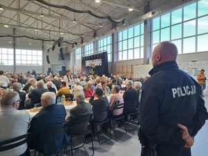 Policjant stoi tyłem do robiącego zdjęcie, a w tle przy stołach siedzą seniorzy.