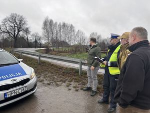 Komisja stoi na poboczu drogi , w tle widoczny jest fragment radiowozu po lewej stronie i w oddali widoczne jest drzewo, w które uderzył samochód renault.