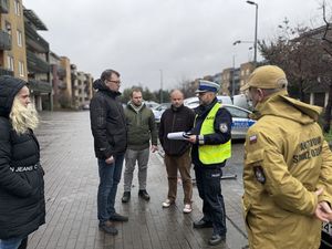 Komisja stoi na drodze przy bloku znajdującym się po lewej stronie a w tle za komisją widać policyjny radiowóz