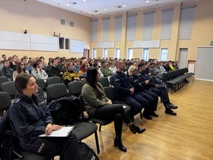 Młodzież wraz z policjantami siedzi na krzesłach, w tym czasie jedna osoba przebywa na scenie i przemawia do publiczności.