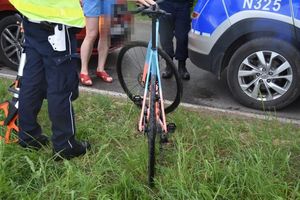 Policjant stoi z rowerem przy radiowozie, w tle stoi jakaś kobieta i widać także obok samochód osobowy