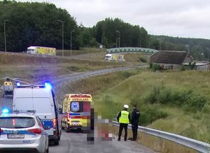 Droga, policyjny radiowóz, za nim w tle śmigłowiec, policjant ruchu drogowego wykonuje czynności służbowe