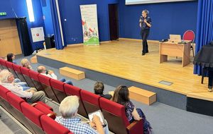 Policjantka prowadzi spotkanie, seniorzy siedzą na widowni w sali konferencyjnej/kinowej.