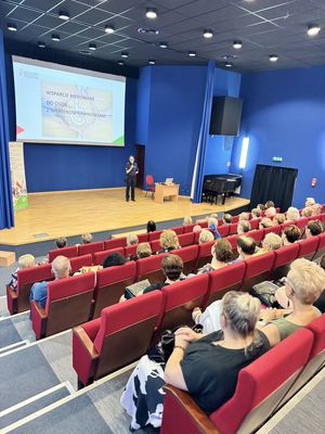 Policjantka prowadzi spotkanie, seniorzy siedzą na widowni w sali konferencyjnej/kinowej.