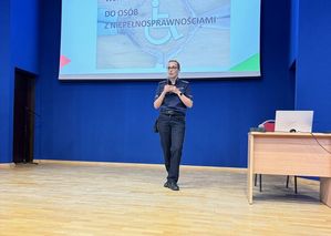 Policjantka prowadzi spotkanie, seniorzy siedzą na widowni w sali konferencyjnej/kinowej.