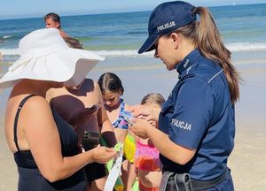 Policjanci  na plaży rozdają opaski niezgubki i ulotki o konkursie