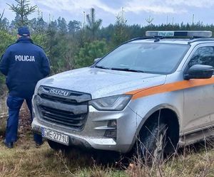 Policjant stoi na linii lasu , w tle stoi radiowóz staży leśnej