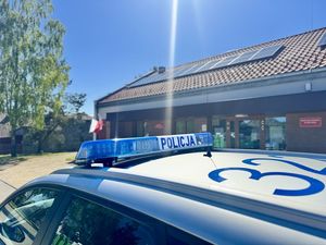 Dach radiowozu z kogutem i w tle widoczny budynek szkoły z flagami biało-czerwonymi .