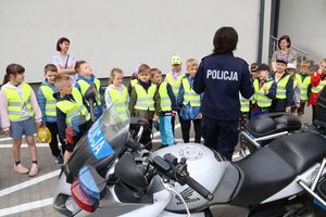 Policjantka prowadzi spotkanie z dziećmi na placu wewnętrznym, na pierwszym planie widoczny jest policyjny motocykl.