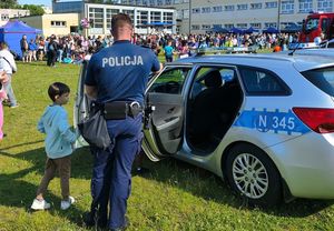 Policjant stoi obok otwartego radiowozu a obok policjanta stoi małe dziecko i widać że coś do niego mówi