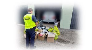 Policjant w kamizelce odblaskowej stoi przed pomieszczeniami w których były składowane towary, które na zdjęciu w części już są na zewnąrz