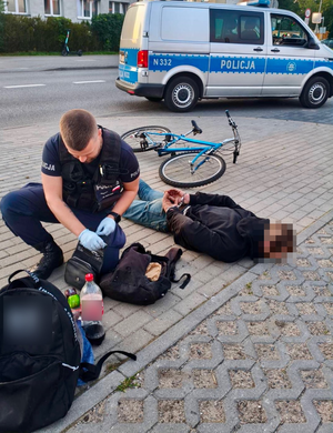 Policjant zakłada kajdanki mężczyźnie leżącemu na chodniku. W tle widać radiowóz oraz leżący rower.