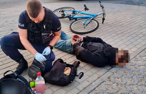 Policjant zakłada kajdanki mężczyźnie leżącemu na chodniku. W tle widać leżący rower.
