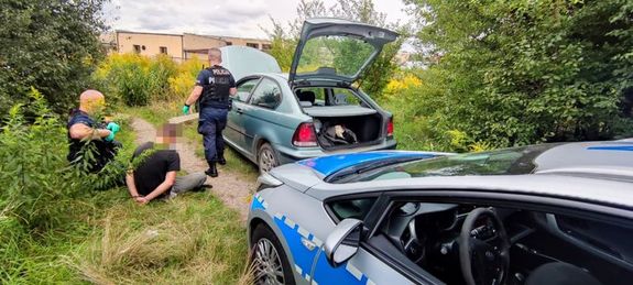 Na zdjęciu jeden z policjantów jest obok zatrzymanego samochodu a drugi wykonuje czynności z zatrzymanym, który siedzi w kajdankach na trawie.
