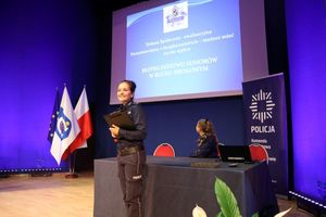Policjantka stoi przy stoliku z komputerem i przez mikrofon przemawia do obecnych gości na sali