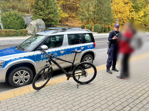 Na zdjęciu stoi radiowóz, obok niebo rower, a po prawej stronie stoi policjantka która rozmawia z mężczyzną.