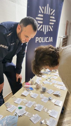 Policjanci rozmawiają z rodzicami i tłumaczą jak niebezpieczne są narkotyki i co przestawiają rekwizyty wystawione na stole.