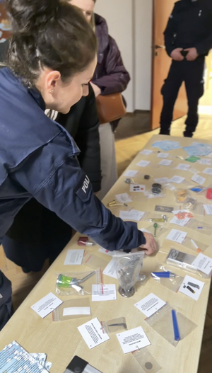 Policjanci rozmawiają z rodzicami i tłumaczą jak niebezpieczne są narkotyki i co przestawiają rekwizyty wystawione na stole.