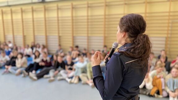 Policjantka prowadzi spotkanie z uczniami na sali. Mówi do nich przez mikrofon.