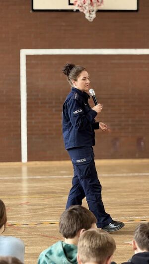 Policjantka prowadzi spotkanie na sali gimnastycznej, dzieci siedzą i słuchają.