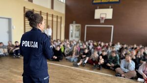 Policjantka prowadzi spotkanie na sali gimnastycznej, dzieci siedzą i słuchają.
