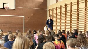 Policjantka prowadzi spotkanie na sali gimnastycznej, dzieci siedzą i słuchają.