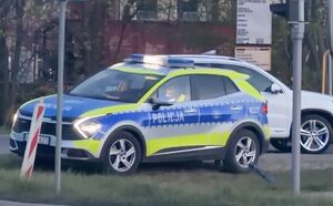 Policyjny radiowóz stoi przy drodze i prowadzi kontrolę drogową pojazdu osobowego w kolorze białym.