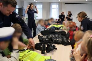 Policjanci oprowadzają dzieci po dziedzińcu komendy, pokazują radiowozy oraz prowadzą spotkanie na sali odpraw, podczas którego dzieci korzystają z zabawek edukacyjnych (samochodów i drogi z przejściem dla pieszych)