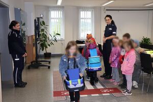 Policjanci oprowadzają dzieci po dziedzińcu komendy, pokazują radiowozy oraz prowadzą spotkanie na sali odpraw, podczas którego dzieci korzystają z zabawek edukacyjnych (samochodów i drogi z przejściem dla pieszych)