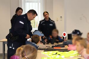 Policjanci oprowadzają dzieci po dziedzińcu komendy, pokazują radiowozy oraz prowadzą spotkanie na sali odpraw, podczas którego dzieci korzystają z zabawek edukacyjnych (samochodów i drogi z przejściem dla pieszych)