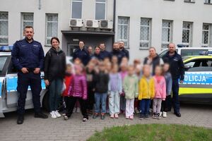 Policjanci oprowadzają dzieci po dziedzińcu komendy, pokazują radiowozy oraz prowadzą spotkanie na sali odpraw, podczas którego dzieci korzystają z zabawek edukacyjnych (samochodów i drogi z przejściem dla pieszych)