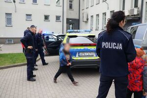 Policjanci oprowadzają dzieci po dziedzińcu komendy, pokazują radiowozy oraz prowadzą spotkanie na sali odpraw, podczas którego dzieci korzystają z zabawek edukacyjnych (samochodów i drogi z przejściem dla pieszych)